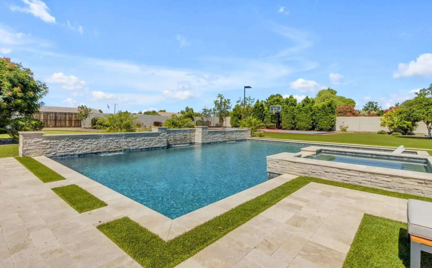 A luxurious backyard features a modern swimming pool with in-pool lounge chairs and a grassy area leading to a basketball court. The scene is bright and sunny, with a clear blue sky and neatly manicured trees and shrubs surrounding the space, all expertly designed by Palm Beach Paving Services