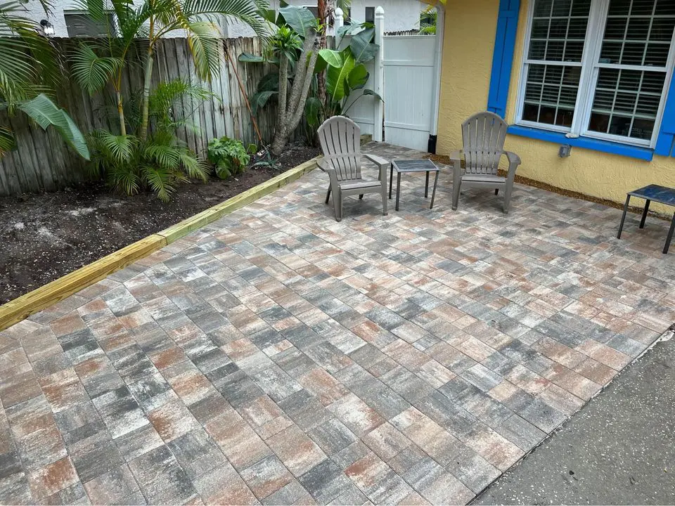 An outdoor patio in Palm Beach County with wicker furniture, white cushions, and colorful pillows. A table holds a tea set with cups, saucers, and pastries. Potted flowers and plants decorate the space, and a curved stone hardscape wall separates the patio from a grassy area.