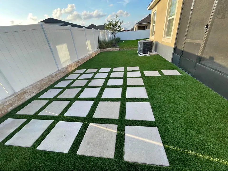 A modern outdoor area in Palm Beach County features a grid pattern of concrete slabs alternated with strips of grass. A palm tree is visible near the corner, and the hardscape appears to be adjacent to a street. The combination of concrete and grass gives a clean and structured look.